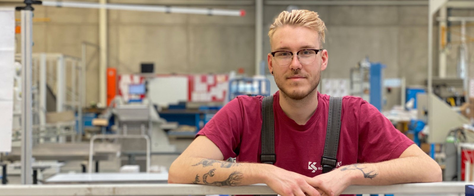Ein junger, blonder Mann in Köberlein & Seigert Arbeitskleidung und mit Brille lächelt in einer Fabrikhalle.